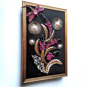 K&ouml;nnte beinhalten: Gerahmtes Kunstwerk mit schwarzem Hintergrund, verziert mit einer Libelle, rosa Edelsteinen, goldenen Halbmondformen und Sternen mit Perlen. Die Anordnung bildet ein florales Design. Der Rahmen ist goldfarben.