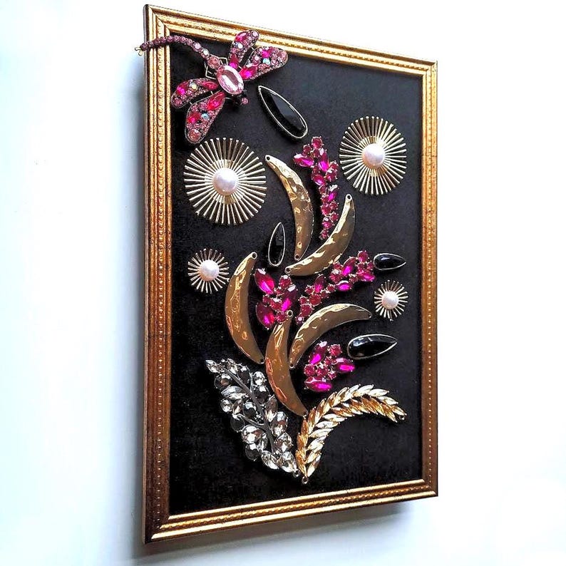 K&ouml;nnte beinhalten: Gerahmtes Kunstwerk mit schwarzem Hintergrund, verziert mit einer Libelle, rosa Edelsteinen, goldenen Halbmondformen und Sternen mit Perlen. Die Anordnung bildet ein florales Design. Der Rahmen ist goldfarben.