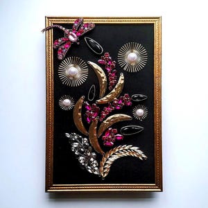 K&ouml;nnte beinhalten: Ein gerahmtes Kunstwerk mit einem schwarzen Hintergrund, verziert mit dekorativen Elementen. Es zeigt goldfarbene Halbmondformen, rosa und klare Edelsteine ​​und eine Libelle. Der Rahmen ist goldfarben.