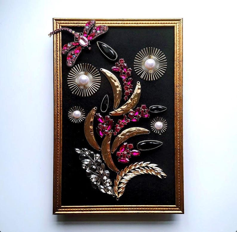 K&ouml;nnte beinhalten: Ein gerahmtes Kunstwerk mit einem schwarzen Hintergrund, verziert mit dekorativen Elementen. Es zeigt goldfarbene Halbmondformen, rosa und klare Edelsteine ​​und eine Libelle. Der Rahmen ist goldfarben.