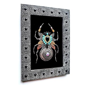 May include: Framed artwork with a decorative spider design. The spider is made from metallic elements, including filigree, beads, and gemstones in black, turquoise, gold, and copper. The silver frame has a black and white pattern and black accents.