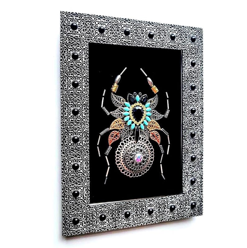May include: Framed artwork with a decorative spider design. The spider is made from metallic elements, including filigree, beads, and gemstones in black, turquoise, gold, and copper. The silver frame has a black and white pattern and black accents.