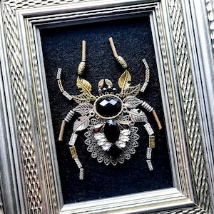 May include: Framed artwork featuring a detailed, mixed-metal spider. The spider has a large, faceted black gemstone body, with silver, gold, and copper-toned metal accents. The artwork is set against a black background within a silver frame.