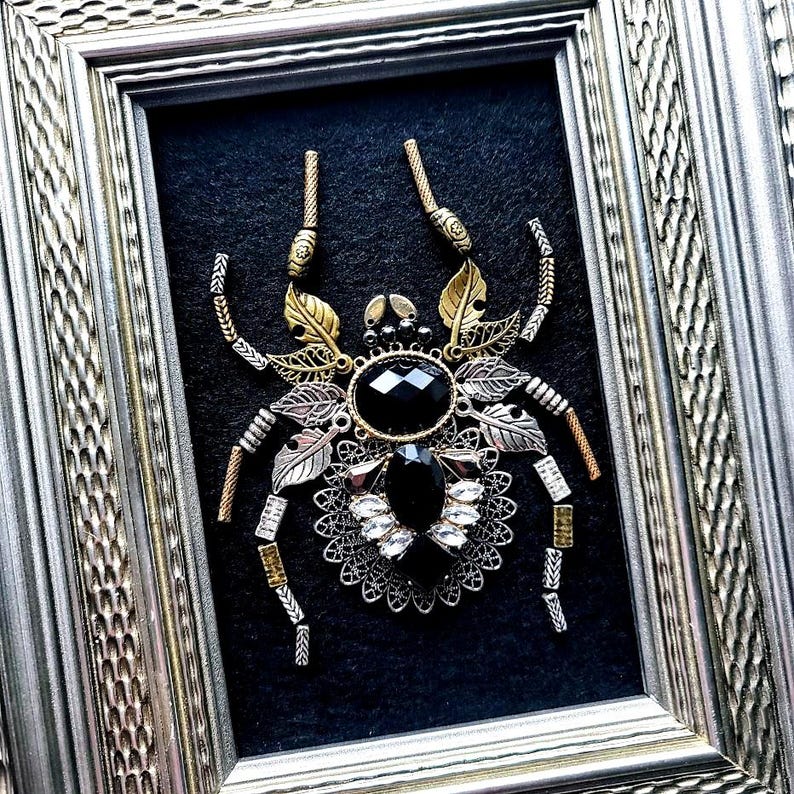 May include: Framed artwork featuring a detailed, mixed-metal spider. The spider has a large, faceted black gemstone body, with silver, gold, and copper-toned metal accents. The artwork is set against a black background within a silver frame.