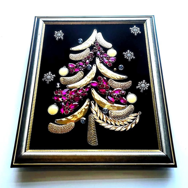 Op de afbeelding: Ingelijst kunstwerk met een kerstboomontwerp. De boom is gemaakt van goudkleurig metaal en versierd met roze edelstenen en witte ornamenten. De achtergrond is zwart en de lijst is goud en zwart.
