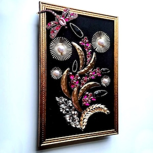 K&ouml;nnte beinhalten: Gerahmtes Kunstwerk mit schwarzem Hintergrund, verziert mit goldenen, rosa und silbernen Verzierungen. Enth&auml;lt eine Libelle, Sonnenstrahlen und florale Elemente. Der Rahmen ist goldfarben.