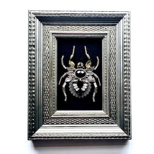May include: A framed, ornate spider sculpture. The spider is crafted from gold and silver-toned metal, with a large black gem at its center. The frame is silver with a textured design, set against a black background.