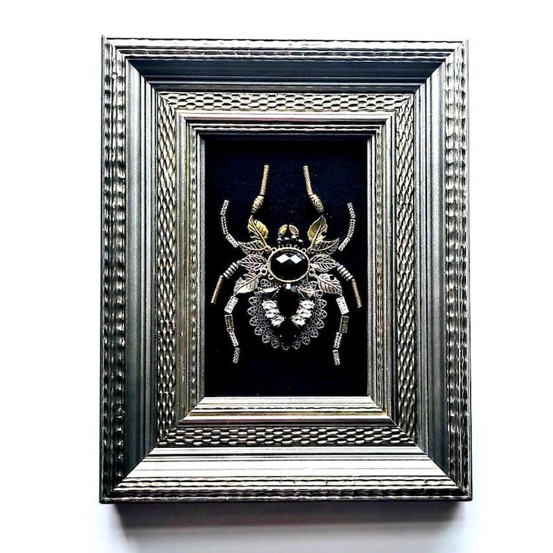 May include: A framed, ornate spider sculpture. The spider is crafted from gold and silver-toned metal, with a large black gem at its center. The frame is silver with a textured design, set against a black background.