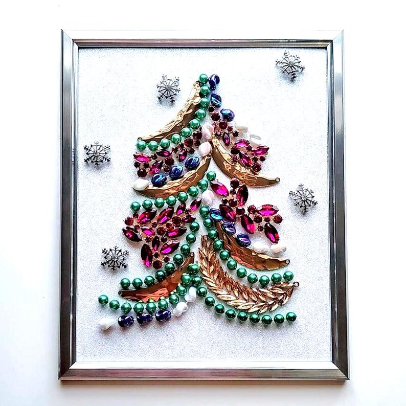 Op de afbeelding: Een ingelijst kerstboomkunstwerk. De boom is gemaakt van groene kralen, roze en blauwe juwelen en goudkleurige metalen bladeren. Zilveren sneeuwvlokversieringen omringen de boom op een witte achtergrond. Het kunstwerk is ingekaderd in een zilveren lijst.