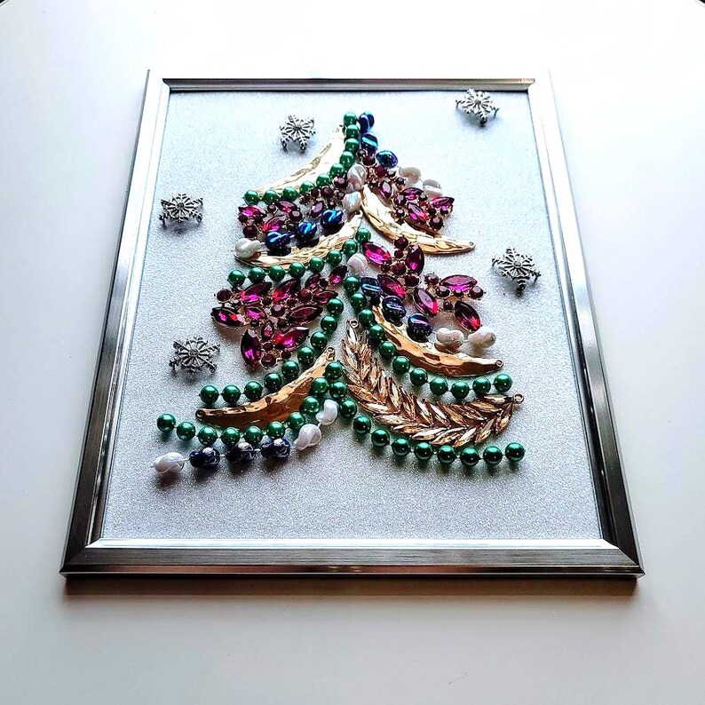Op de afbeelding: Een ingelijst kerstboomkunstwerk. De boom is gemaakt met groene, witte en paarse kralen en goudkleurige bladeren. Zilveren sneeuwvlokken zijn verspreid over een glitterachtergrond. De lijst is zilver.
