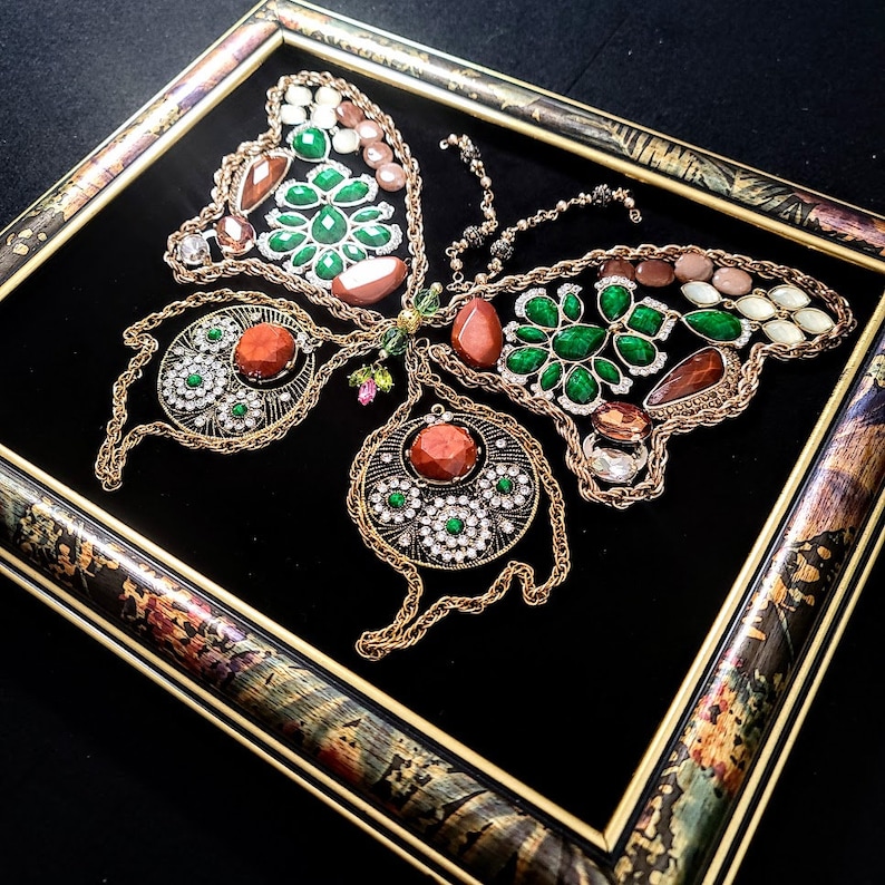 K&ouml;nnte beinhalten: Ein gerahmtes Schmetterlingskunstwerk aus goldenen Ketten und verschiedenen farbigen Edelsteinen, darunter gr&uuml;n, rot und braun. Der Schmetterling ist vor einem schwarzen Samt Hintergrund platziert.