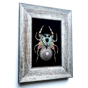 May include: Framed artwork depicting a spider design. The spider is made from silver, gold, and turquoise metallic elements, set against a black backdrop. The frame is a distressed grey wood.