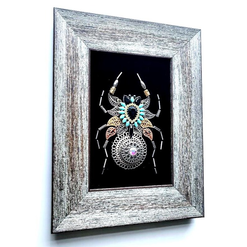 May include: Framed artwork depicting a spider design. The spider is made from silver, gold, and turquoise metallic elements, set against a black backdrop. The frame is a distressed grey wood.