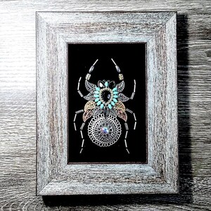 May include: Framed artwork with a decorative spider design. The spider is made from silver, gold, and turquoise metallic elements, set against a black background. The frame is a distressed, light grey wood.