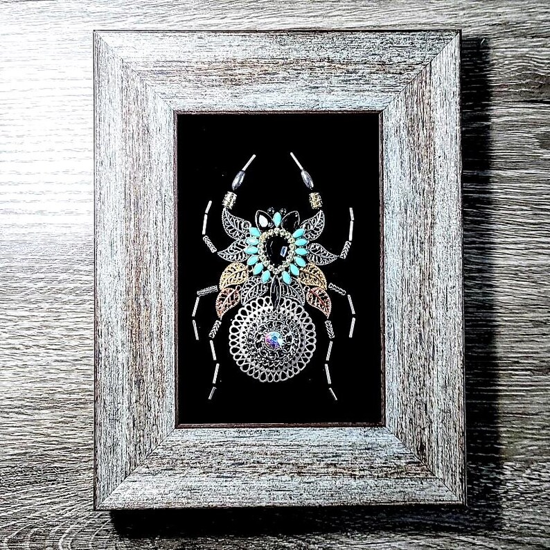May include: Framed artwork with a decorative spider design. The spider is made from silver, gold, and turquoise metallic elements, set against a black background. The frame is a distressed, light grey wood.