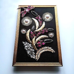 K&ouml;nnte beinhalten: Ein gerahmtes Kunstwerk mit schwarzem Hintergrund, verziert mit goldfarbenen Halbmonden, rubinroten Kristallclustern und schwarzen Tropfenakzenten. Eine Libelle und Blattakzente vervollst&auml;ndigen das florale Design. Der Rahmen hat eine goldene Oberfl&auml;che.