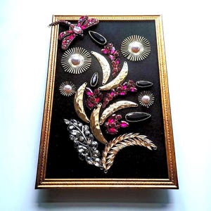 K&ouml;nnte beinhalten: Ein gerahmtes Kunstwerk mit einer Anordnung von Vintage-Broschen und Verzierungen. Die Komposition umfasst eine Libelle, Halbmondformen und florale Elemente in Gold-, Silber- und Rubinrott&ouml;nen auf schwarzem Hintergrund.
