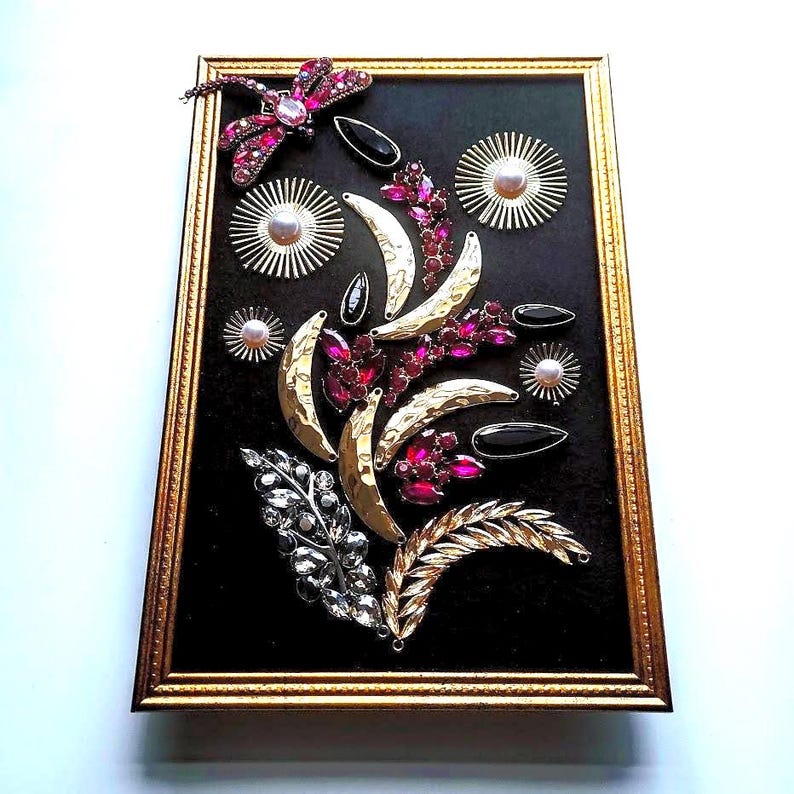 K&ouml;nnte beinhalten: Ein gerahmtes Kunstwerk mit einer Anordnung von Vintage-Broschen und Verzierungen. Die Komposition umfasst eine Libelle, Halbmondformen und florale Elemente in Gold-, Silber- und Rubinrott&ouml;nen auf schwarzem Hintergrund.