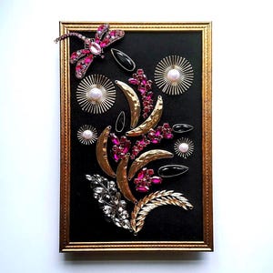 K&ouml;nnte beinhalten: Ein gerahmtes Kunstwerk mit schwarzem Hintergrund, verziert mit goldfarbenen Halbmonden, rosa und klaren Edelsteinen und einer Libelle. Der Rahmen ist goldfarben. Das Design hat ein florales Thema.