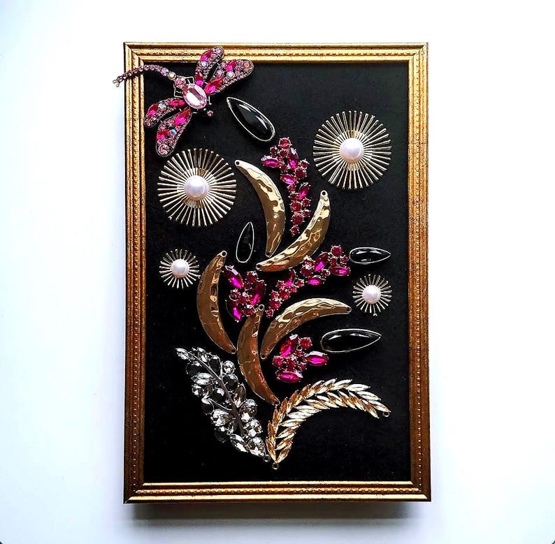 K&ouml;nnte beinhalten: Ein gerahmtes Kunstwerk mit schwarzem Hintergrund, verziert mit goldfarbenen Halbmonden, rosa und klaren Edelsteinen und einer Libelle. Der Rahmen ist goldfarben. Das Design hat ein florales Thema.
