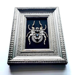 May include: Framed artwork featuring a detailed spider design. The spider is crafted from metallic elements, with a large black gemstone at its center. The artwork is set against a black background within a silver-toned frame.