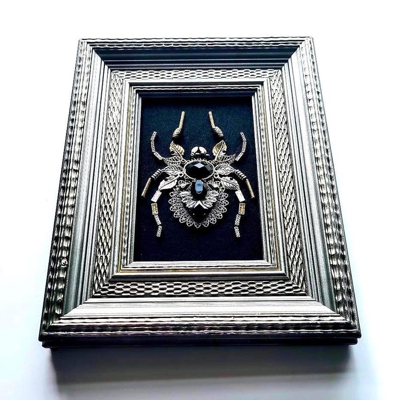 May include: Framed artwork featuring a detailed spider design. The spider is crafted from metallic elements, with a large black gemstone at its center. The artwork is set against a black background within a silver-toned frame.