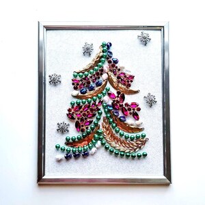 Op de afbeelding: Een ingelijst kerstboomkunstwerk. De boom is gemaakt van diverse kleurrijke juwelen, waaronder groene kralen, roze en paarse edelstenen en goudkleurige bladeren. Zilveren sneeuwvlokken zijn verspreid rond de boom op een witte achtergrond.