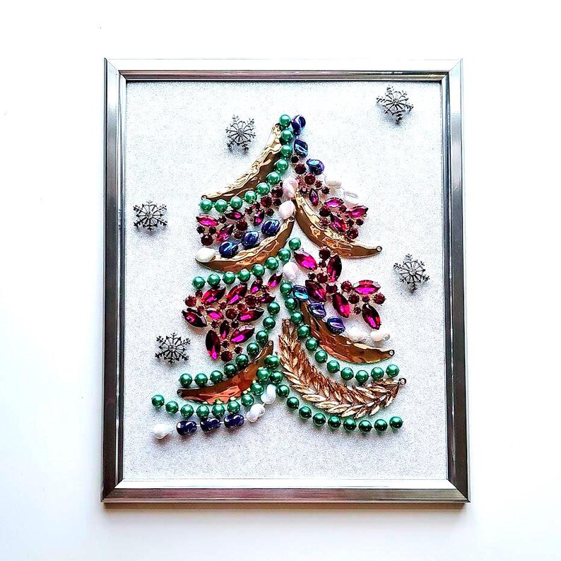 Op de afbeelding: Een ingelijst kerstboomkunstwerk. De boom is gemaakt van diverse kleurrijke juwelen, waaronder groene kralen, roze en paarse edelstenen en goudkleurige bladeren. Zilveren sneeuwvlokken zijn verspreid rond de boom op een witte achtergrond.