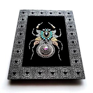 May include: A framed artwork with a jeweled spider design on a black background. The spider is made with turquoise, gold, and silver-toned embellishments. The frame is black and white with a heart and swirl pattern.