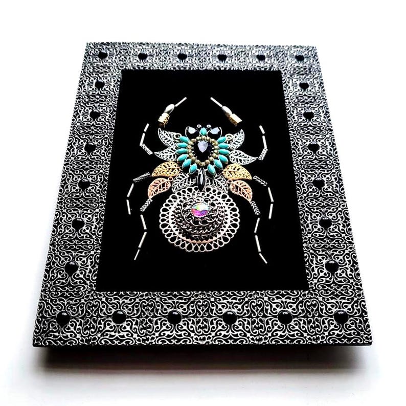 May include: A framed artwork with a jeweled spider design on a black background. The spider is made with turquoise, gold, and silver-toned embellishments. The frame is black and white with a heart and swirl pattern.