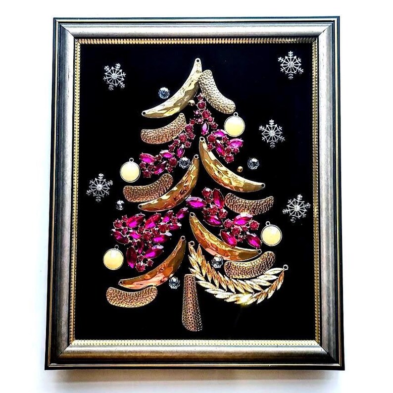 Op de afbeelding: Ingelijst kerstboomkunstwerk. De boom is gemaakt van goudkleurige en getextureerde elementen, roze juwelen en witte ornamenten, tegen een zwarte achtergrond. Zilveren sneeuwvlokken en een zilveren lijst maken het feestelijke ontwerp compleet.
