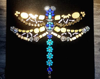May include: A dragonfly made of colorful rhinestones and beads on a black background. The dragonfly's wings are made of yellow, white, and purple rhinestones. The body is made of blue and turquoise rhinestones.
