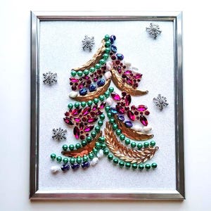 Op de afbeelding: Een ingelijst kerstboomkunstwerk. De boom is gemaakt van kleurrijke juwelen, kralen en goudkleurige bladvormen. De achtergrond is een glinsterend wit, met zilveren sneeuwvlokaccenten.