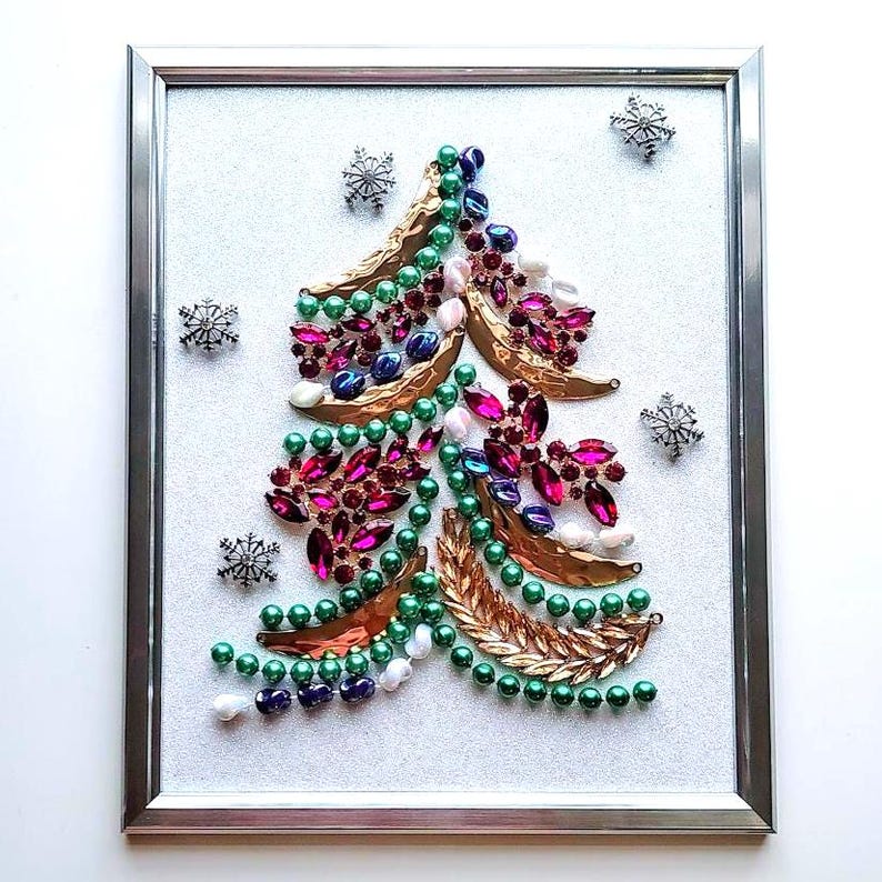 Op de afbeelding: Een ingelijst kerstboomkunstwerk. De boom is gemaakt van kleurrijke juwelen, kralen en goudkleurige bladvormen. De achtergrond is een glinsterend wit, met zilveren sneeuwvlokaccenten.
