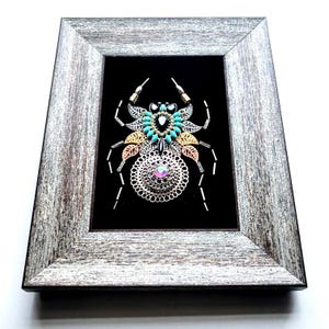 May include: A framed artwork depicting a spider constructed from metal and beads. The spider's body incorporates silver, gold, and copper elements, with turquoise beads and an iridescent gem. It's set against a black backdrop within a gray, rustic wooden frame.