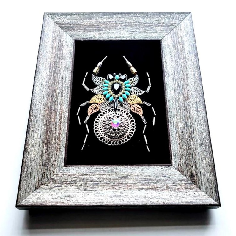May include: A framed artwork depicting a spider constructed from metal and beads. The spider's body incorporates silver, gold, and copper elements, with turquoise beads and an iridescent gem. It's set against a black backdrop within a gray, rustic wooden frame.