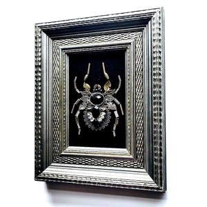 May include: A framed artwork featuring a detailed spider design. The spider is crafted with metallic elements, including gold and silver accents, and a large black gemstone at its center. The artwork is set against a black background within a silver frame.