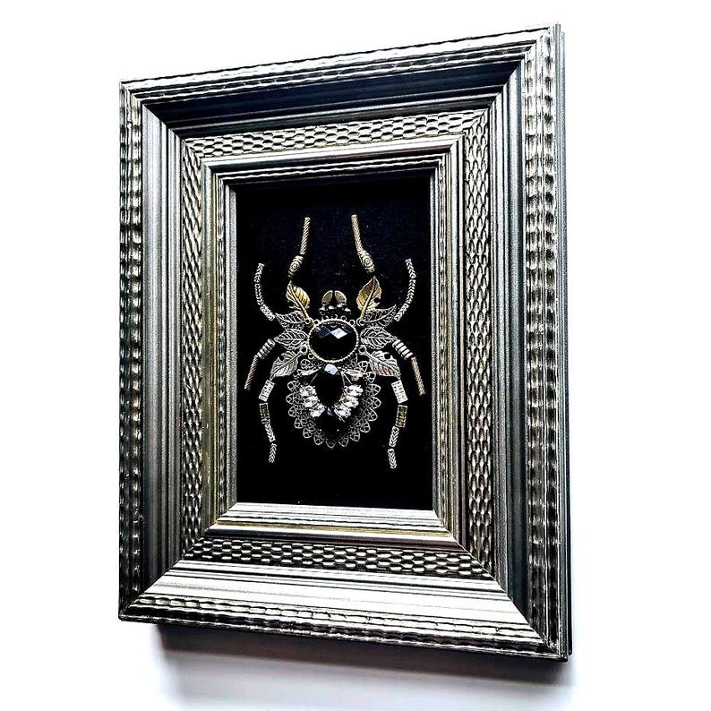 May include: A framed artwork featuring a detailed spider design. The spider is crafted with metallic elements, including gold and silver accents, and a large black gemstone at its center. The artwork is set against a black background within a silver frame.