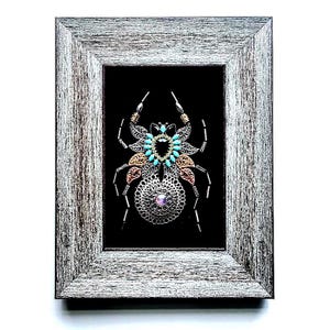May include: Framed artwork of a metal spider with turquoise, gold, and silver detailing, and a central pink gem. The distressed grey wooden frame surrounds a black background. The artwork is a decorative piece.