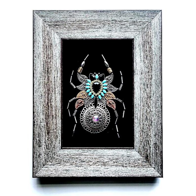 May include: Framed artwork of a metal spider with turquoise, gold, and silver detailing, and a central pink gem. The distressed grey wooden frame surrounds a black background. The artwork is a decorative piece.
