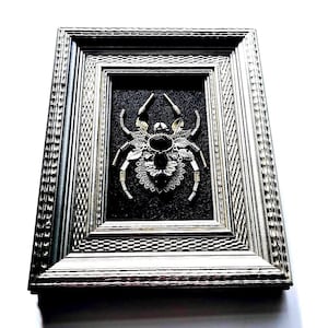May include: Framed artwork featuring a detailed spider design. The spider is crafted from silver and gold-toned metal components, with a large black gemstone at its center. The artwork is set against a dark background within a silver frame.