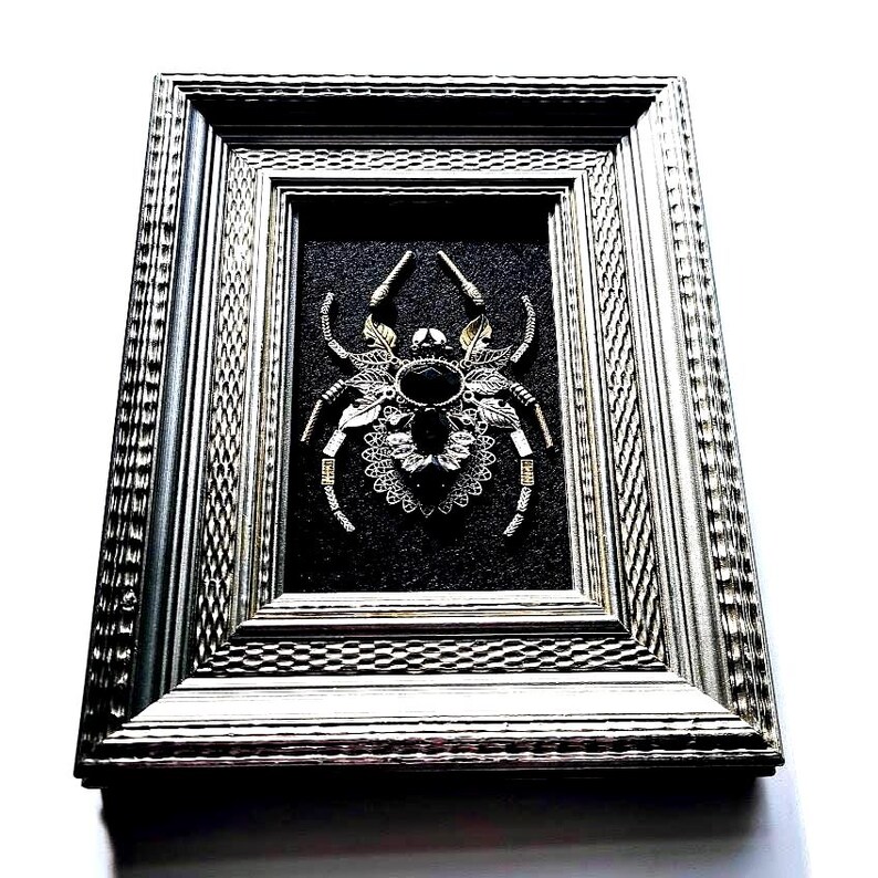 May include: Framed artwork featuring a detailed spider design. The spider is crafted from silver and gold-toned metal components, with a large black gemstone at its center. The artwork is set against a dark background within a silver frame.