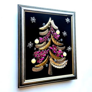 Op de afbeelding: Ingelijst kunstwerk met een kerstboomontwerp. De boom is samengesteld uit goudkleurige banaanvormige elementen, roze juwelen en decoratieve accenten. De achtergrond is zwart en de lijst is zilver. Sneeuwvlok- en ornamentdetails maken de feestelijke look compleet.