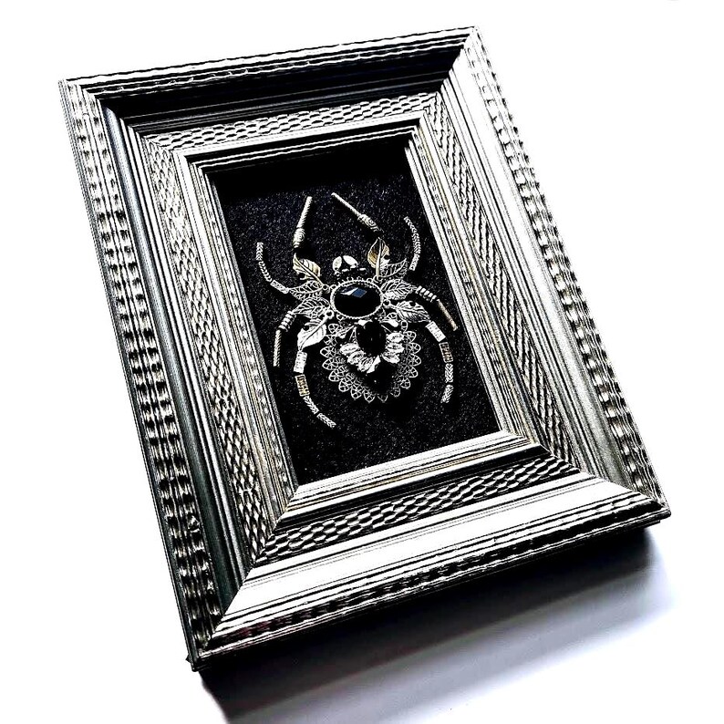 May include: A framed, silver-toned spider brooch with a large black gemstone center. The spider is intricately detailed with delicate legs and decorative accents. The brooch is set against a dark gray background within a silver frame, creating a striking contrast.