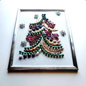Op de afbeelding: Een ingelijst kerstboomkunstwerk. De boom is gemaakt van groene, paarse, blauwe en witte kralen en edelstenen, met gouden bladaccenten. Zilveren sneeuwvlokken zijn verspreid rond de boom op een glitterachtergrond.