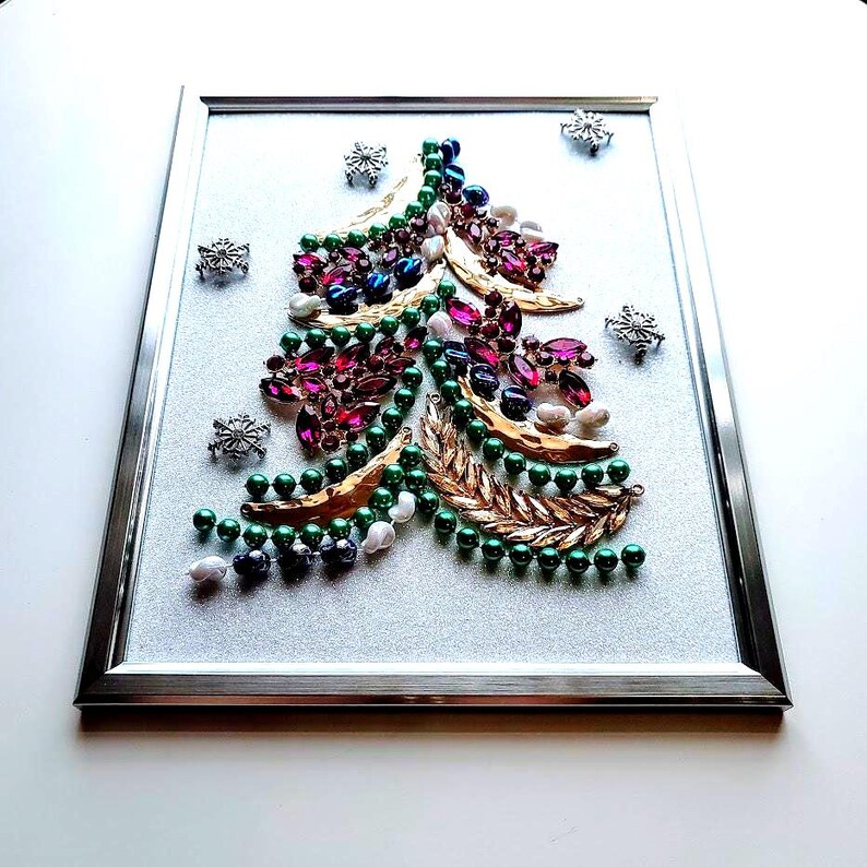 Op de afbeelding: Een ingelijst kerstboomkunstwerk. De boom is gemaakt van groene, paarse, blauwe en witte kralen en edelstenen, met gouden bladaccenten. Zilveren sneeuwvlokken zijn verspreid rond de boom op een glitterachtergrond.