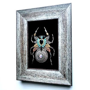 May include: Framed artwork of a decorative spider, crafted from metal elements and turquoise beads, set against a black backdrop. The rustic grey wooden frame surrounds the spider. A unique, handcrafted piece of art.