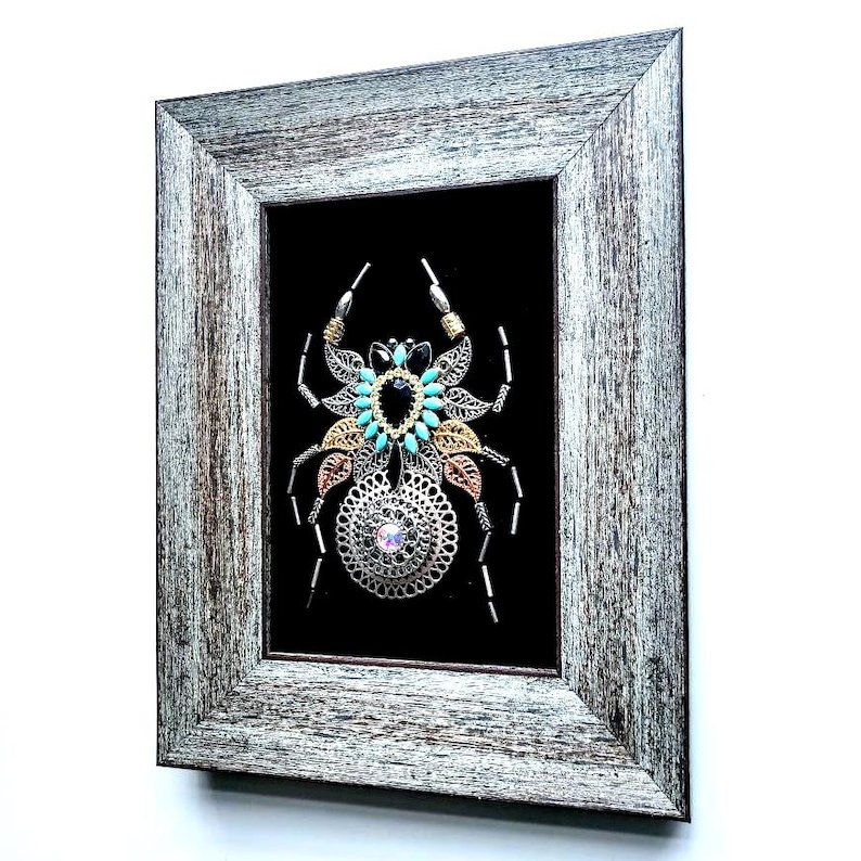 May include: Framed artwork of a decorative spider, crafted from metal elements and turquoise beads, set against a black backdrop. The rustic grey wooden frame surrounds the spider. A unique, handcrafted piece of art.
