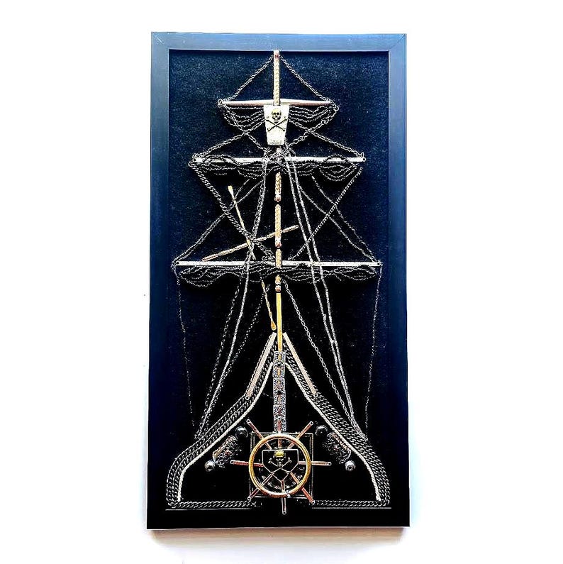 Pode incluir: Uma obra de arte emoldurada que retrata um mastro de navio pirata, feito de correntes de metal e detalhes. O navio apresenta uma bandeira de caveira e ossos cruzados, um leme dourado e uma aparelhagem intrincada contra um fundo azul escuro. A moldura &eacute; preta.