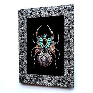 May include: Framed artwork of a spider, crafted from metal components. The spider features silver, gold, and copper elements, with turquoise accents and a central iridescent gem. The silver frame has a black pattern and is studded with black beads.